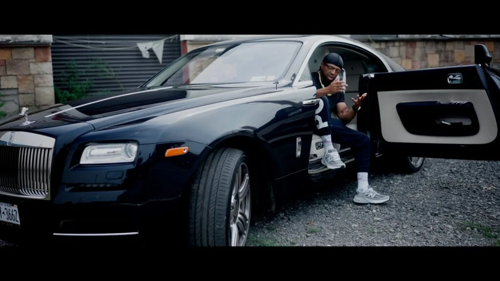 Music video still featuring a performer beside a Rolls-Royce